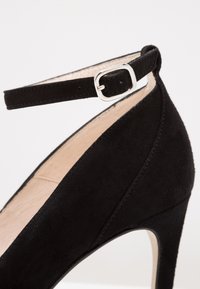 Chaussure à talon haut en daim noir avec une bride à la cheville, ornée d'une boucle argentée. La chaussure a un bout pointu et une doublure intérieure lisse.