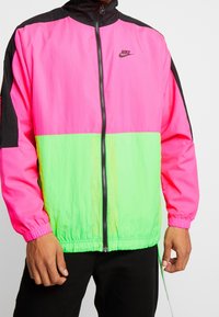 Person som bär en ljusrosa och neongrön Nike zip-up jacka med svarta detaljer och svarta byxor, händerna avslappnat vid sidorna.