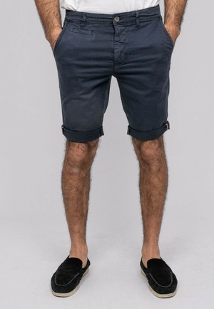 Homme portant un short bleu marine foncé retroussé, une chemise blanche, des chaussures noires à enfiler, debout les mains dans les poches devant un fond neutre.