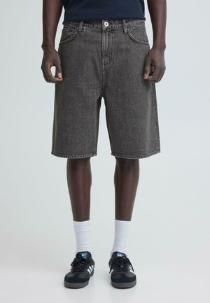 Ciemnoszare szorty jeansowe noszone przez osobę stojącą, zestawione z białymi skarpetkami i czarnymi sneakersami Adidas z białymi paskami.