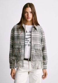 Ruudullinen, fringed takki harmaissa ja vaaleanpunaisissa sävyissä, jossa on kauluksellinen kaula-aukko ja kaksi etutaskua. Vaatteena valkoinen graafinen t-paita.