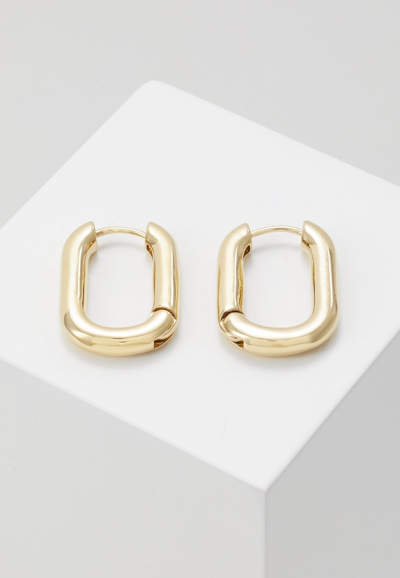 Paire de boucles d’oreilles créoles rectangulaires dorées brillantes avec des bords arrondis présentées sur un socle blanc sur fond gris clair.