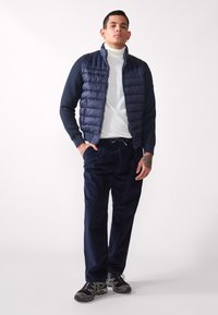 Marinblå pufferväst med kviltad struktur över en vit polotröja, kombinerad med mörka corduroybyxor och svarta sportskor. Avslappnad outfit.