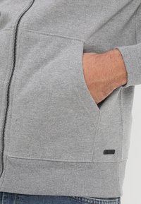 Grå sweatshirt med lynlås og ribbede manchetter, med sidelommer og en lille sort labeldetalje. Blødt stof med en tekstureret overflade.