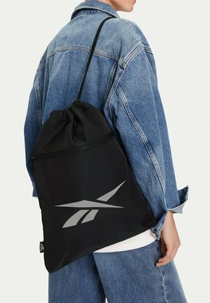 Persona con chaqueta de mezclilla y vaqueros que lleva una bolsa negra con cordón y un logo plateado de Reebok en la espalda.