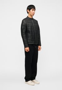 Μαύρο δερμάτινο μπουφάν με καπιτονέ σχέδιο, όρθιο γιακά, πλαϊνά φερμουάρ και δομημένη σχεδίαση, συνδυασμένο με άνετο μαύρο παντελόνι και λευκά αθλητικά παπούτσια.