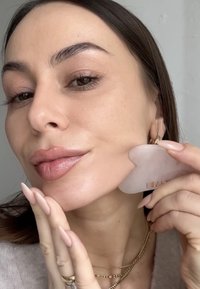 Un outil facial translucide en forme de cœur est tenu contre une joue lisse de couleur beige clair. La personne a des ongles longs de teinte neutre et des bijoux en or superposés.