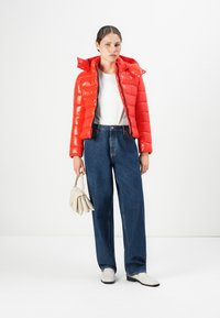 Vrouw die staat met een felrode pufferjack, witte shirt, blauwe jeans, witte schoenen, en een witte handtas tegen een witte achtergrond.
