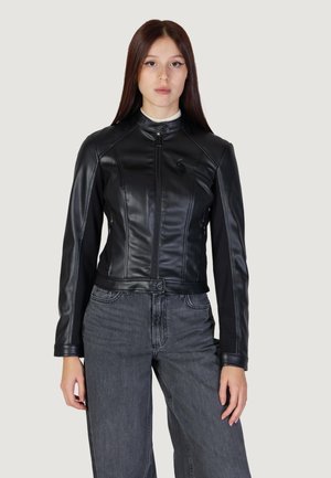 Giacca di pelle nera con un design aderente, chiusura a zip e una piccola tasca sul petto. Abbinata a jeans grigi a gamba larga e un dolcevita bianco.