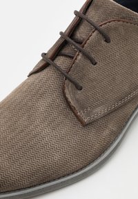 Scarpa in suede marrone con lacci, caratterizzata da un motivo a spina di pesce, lacci marroni e una suola in gomma grigia. Texture liscia con cuciture dettagliate.