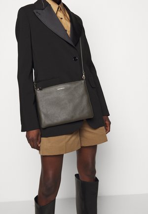 Osoba o ciemnej karnacji ubrana w czarną marynarkę, beżowe szorty, czarne kozaki do kolan oraz nosząca ciemnoszarą skórzaną torebkę na ramię zapinaną na zamek.