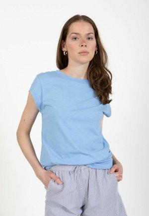 Giovane donna con capelli castani lunghi che indossa una maglietta azzurro chiaro a maniche corte e pantaloni a righe con coulisse, mani nelle tasche, espressione neutra.
