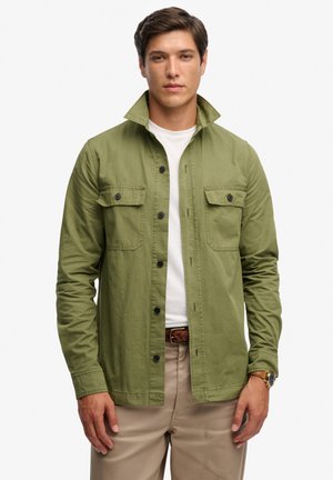 Mężczyzna w oliwkowo-zielonej koszuli na guziki, założonej na białą koszulkę, beżowe spodnie, brązowy pleciony pasek oraz zegarek na rękę na jednolitym białym tle.