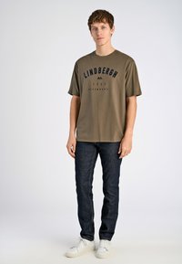 Camiseta verde oliva con texto azul marino que dice "LINDBERGH 1921 COPENHAGUE", combinada con jeans oscuros y zapatillas blancas. Mangas cortas, corte relajado.