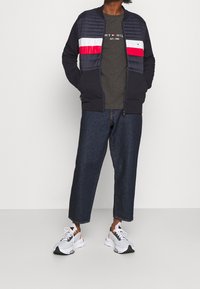 Námornícká bomber bunda s prošívaným předkem a červenobílými pruhovanými detaily. Nositá přes šedé tričko, doplněná tmavými džíny a bílými teniskami.