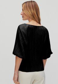 Sort plisseret top med rund halsudskæring, pufærmer og tekstureret stof. Båret med lyse bukser, der viser tilbageperspektivet.