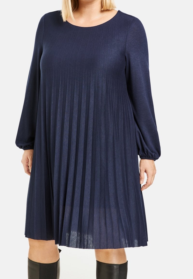 Femme portant une robe bleu marine à manches longues, plissée et longueur genou, avec un col rond, et des bottes noires montantes jusqu'aux genoux.