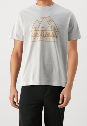 Man wearing white short-sleeve t-shirt with brown mountain, trees, tent graphic and "NAPAPIJRI geographic" text on front.