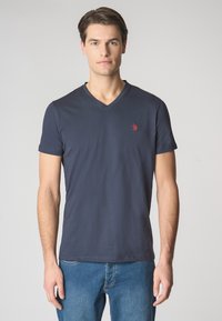 T-shirt a V blu realizzata in cotone, con maniche corte e un piccolo logo rosso sul lato sinistro del petto. Indossata con jeans blu.