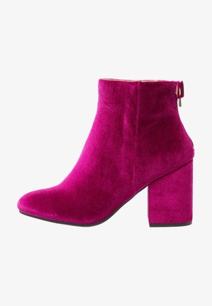 Bota de terciopelo fucsia hasta el tobillo con tacón ancho y cremallera lateral, mostrando el perfil izquierdo contra un fondo blanco.
