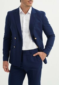 Blazer croisé bleu marine avec des boutons dorés, associé à une chemise blanche à boutons. Coupe ajustée, épaules structurées et poche avant.