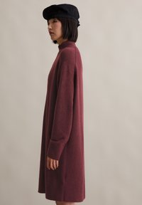 Frau in einem langen, kastanienbraunen Pulloverkleid und einer schwarzen Baskenmütze, die im Profil vor einem schlichten beigen Hintergrund steht.