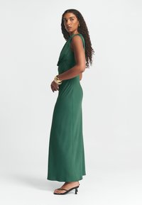 Zelené saténové maxi šaty s drapovaným výstřihem, bez rukávů a přiléhavým střihem. Nosí se s černými sandály na podpatku.