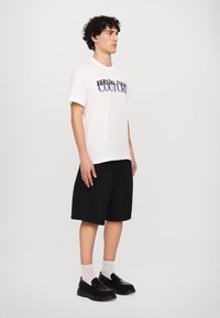 Biely tričko s krátkym rukávom a fialovým nápisom "VERSACE COUTURE", skombinované s čiernymi šortkami a masívnymi čiernymi nazúvacími topánkami.