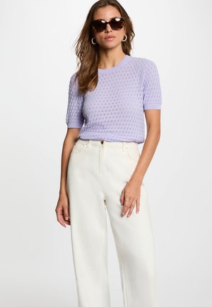 Frau mit Schildpatt-Sonnenbrille, goldenen Creolen, einem kurzärmeligen fliederfarbenen Stricktop und hoch taillierten weißen weiten Hosen.