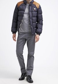 Dunkelblauer Steppmantel mit senffarbenen Lederakzenten an den Schultern, Reißverschluss und Logoaufnäher, kombiniert mit hellgrauer Hose und schwarzen Schuhen.