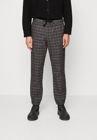 Karierte Hose aus dunklem Stoff mit hellem Gittermuster, einem elastischen Bund mit Kordelzug und schmal zulaufenden Bündchen. Wird mit schwarzen Stiefeln getragen.