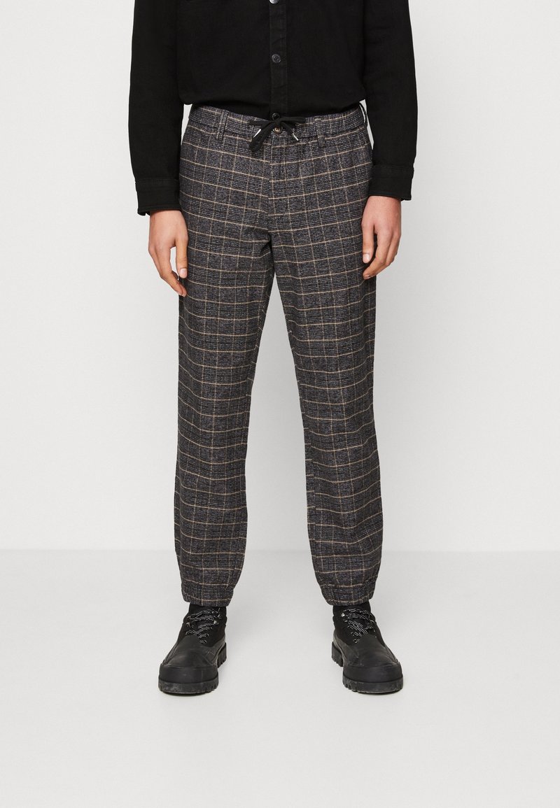 Karierte Hose aus dunklem Stoff mit hellem Gittermuster, einem elastischen Bund mit Kordelzug und schmal zulaufenden Bündchen. Wird mit schwarzen Stiefeln getragen.