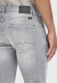 Jean en denim gris avec une coupe droite, des coutures visibles, un patch logo sur la ceinture et une forme de poche arrière unique.