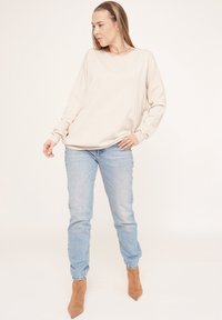 Hellbeiges, oversized Sweater mit überschnittenen Schultern, kombiniert mit hellblauen Jeans und tanfarbenen, spitz zulaufenden Absätzen, vor einem neutralen Hintergrund.