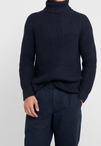 Homme portant un pull à col roulé en maille épaisse bleu marine et un pantalon ample assorti, une main dans la poche.