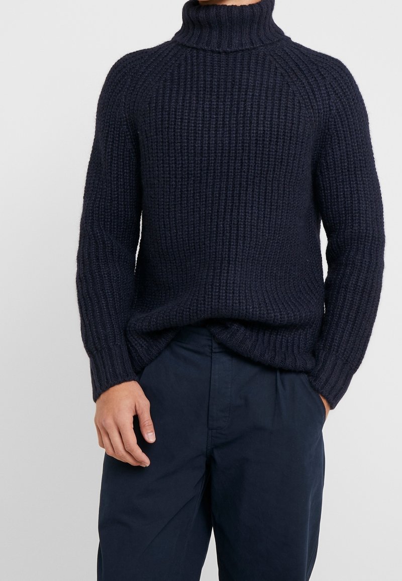 Homme portant un pull à col roulé en maille épaisse bleu marine et un pantalon ample assorti, une main dans la poche.