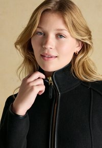 Junge Frau mit blondem Haar, die eine schwarze Jacke mit Reißverschluss trägt und den Reißverschluss nahe am Kinn hält, vor einem einfarbigen beigen Hintergrund.