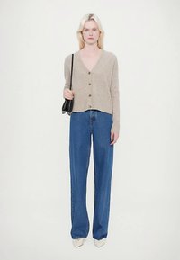 Bézs színű kötött kardigán V-nyakú és gombos záródással, kényelmes szabású kék farmernadrággal, és hegyes orrú, fehér magassarkú cipővel párosítva.