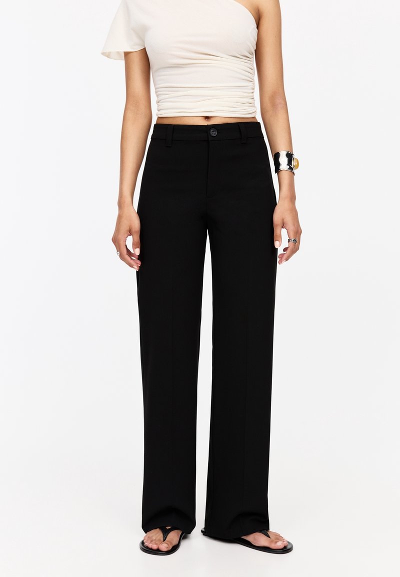 Pantaloni neri a gamba larga con chiusura a bottone, tessuto liscio e design sartoriale. Abbinati a una maglietta aderente color crema e infradito.
