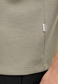 Beeži vahvlilise kootud kangaga särk, mille ääres on väike valge "Jack & Jones" silt, kombineeritud mustade pükstega ja nähtav osa käest.