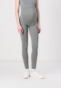 Leggings de maternidade em um tecido verde suave e apagado. Design ajustado com uma textura lisa e sem padrões visíveis. Combinados com tênis brancos.