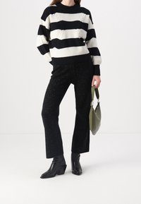 Suéter de punto a rayas en blanco y negro con patrón de cable, combinado con pantalones rectos negros brillantes y botas de tobillo negras de tacón, llevando un bolso verde.