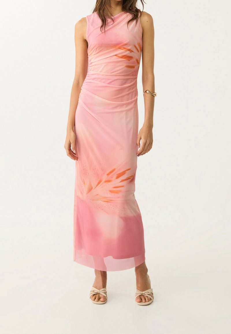 Femme portant une robe longue rose sans manches avec des motifs floraux orange, des talons beiges ouverts à l'avant et un bracelet en or au bras droit.
