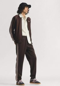 Brązowy dres z białymi akcentami, zawierający kurtkę na zamek, spodnie z elastycznym paskiem oraz różowe trampki w paski z brązową podeszwą.