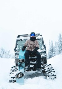 Snowboard mit blauem und schwarzem Design, Camouflage-Bindungen, liegt auf einem schneebedeckten Fahrzeug mit robustem Design in einer verschneiten Landschaft.
