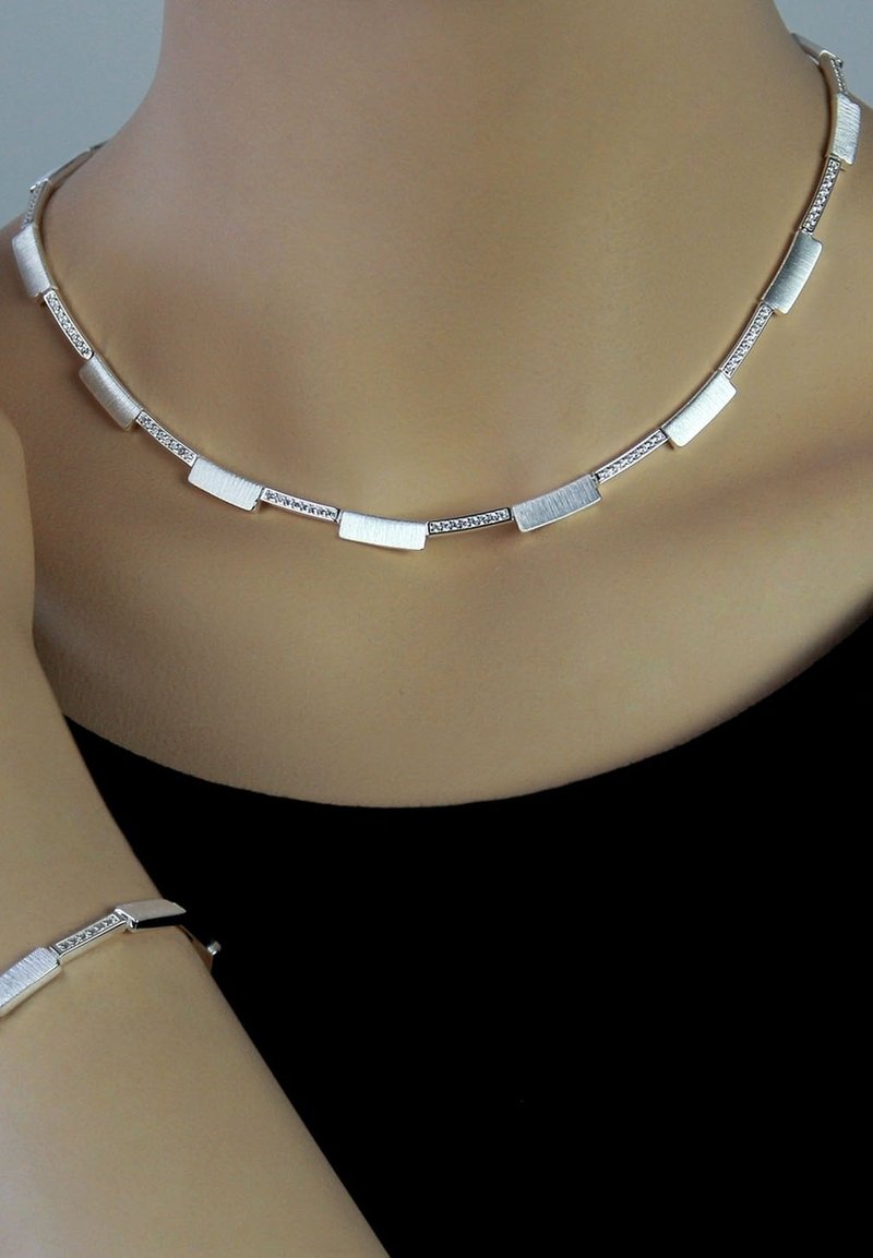 Collier en argent composé de maillons rectangulaires texturés et de petits accents ronds, porté autour du cou, avec un bracelet assorti visible.