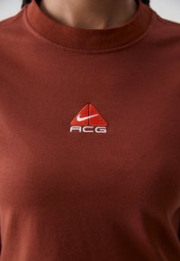 Brown long-sleeve shirt featuring a ribbed crew neck. Centered embroidered logo with red triangle and white "ACG" and swoosh accents.