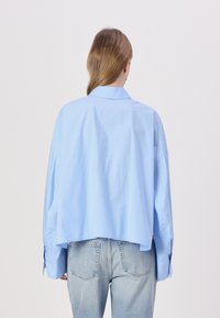 Soehhelblue värvi ülev suuruses särk, millel on krae ja pikad varrukad, kastikujuline lõige ja nööpidega mansetid, sobitatu pleekinud denim pükstega.
