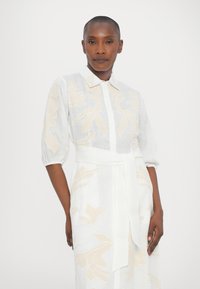 White linen shirt dress with puffed sleeves, featuring a floral pattern in beige. Includes a sash belt and a collared neckline.