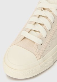 Béžová teniska s krémovou gumovou špičkou. Má měkký semišový svršek, krémové tkaničky a texturovaný vzor na podrážce.